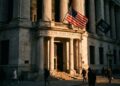 Fotografía documental de la fachada de la Bolsa de Nueva York (NYSE) al atardecer. La luz dorada baja proyecta sombras largas y dramáticas sobre las columnas corintias y la entrada principal, mientras la bandera estadounidense ondea al viento. Peatones con ropa de invierno caminan en primer plano bajo una atmósfera solemne.