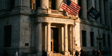 Fotografía documental de la fachada de la Bolsa de Nueva York (NYSE) al atardecer. La luz dorada baja proyecta sombras largas y dramáticas sobre las columnas corintias y la entrada principal, mientras la bandera estadounidense ondea al viento. Peatones con ropa de invierno caminan en primer plano bajo una atmósfera solemne.