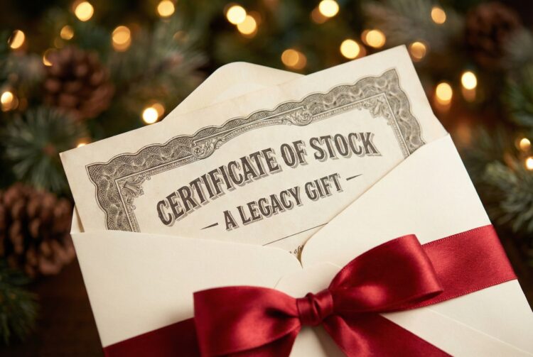 Fotografía de primer plano que muestra un certificado de acciones de estilo vintage con bordes grabados y el texto "CERTIFICATE OF STOCK - A LEGACY GIFT", emergiendo de un sobre de regalo color crema atado con un elegante lazo de cinta de satén roja. El fondo es un desenfoque cálido (bokeh) de luces navideñas doradas, ramas de pino y piñas, sugiriendo un regalo de valor duradero.