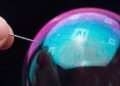 Fotografía macro conceptual que muestra una aguja plateada, sostenida por una mano, tocando la superficie de una gran burbuja de jabón iridiscente. La burbuja refleja patrones brillantes de circuitos digitales azules y el texto "AI" en el centro, sobre un fondo negro.