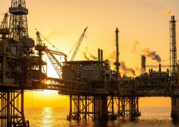 Fotografía panorámica al amanecer de una gran plataforma petrolera marina y complejo de refinería. Las complejas estructuras metálicas, grúas y columnas de humo se siluetean contra un cielo naranja y dorado brillante sobre el océano. El logotipo de "EXXON" es claramente visible en varias partes de la infraestructura, simbolizando la actividad y la oportunidad en el sector energético.