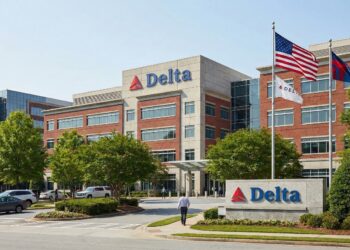 Fotografía exterior diurna de la sede corporativa de Delta Air Lines en Atlanta bajo un cielo soleado. El edificio moderno de ladrillo y hormigón presenta el logotipo triangular rojo y el texto azul "Delta" en la parte superior de la fachada y en un letrero de monumento de piedra cerca de la entrada a nivel de calle. Las banderas de Estados Unidos, una blanca con el logo de Delta y otra azul ondean en mástiles altos. El entorno está cuidadosamente ajardinado con árboles verdes, y una persona camina por la acera hacia el edificio.