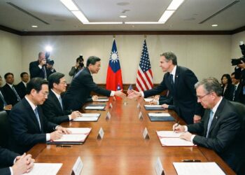 Una fotografía de una sala de conferencias formal muestra a un alto funcionario estadounidense (a la derecha, parecido a Antony Blinken) estrechando la mano de un alto funcionario taiwanés (a la izquierda) sobre una larga mesa de madera pulida. Ambos han estado firmando documentos abiertos frente a ellos. Las banderas de Estados Unidos y de la República de China (Taiwán) están colocadas verticalmente detrás de ellos. Otros funcionarios observan sentados a la mesa, mientras fotógrafos con cámaras y flashes trabajan en el fondo.