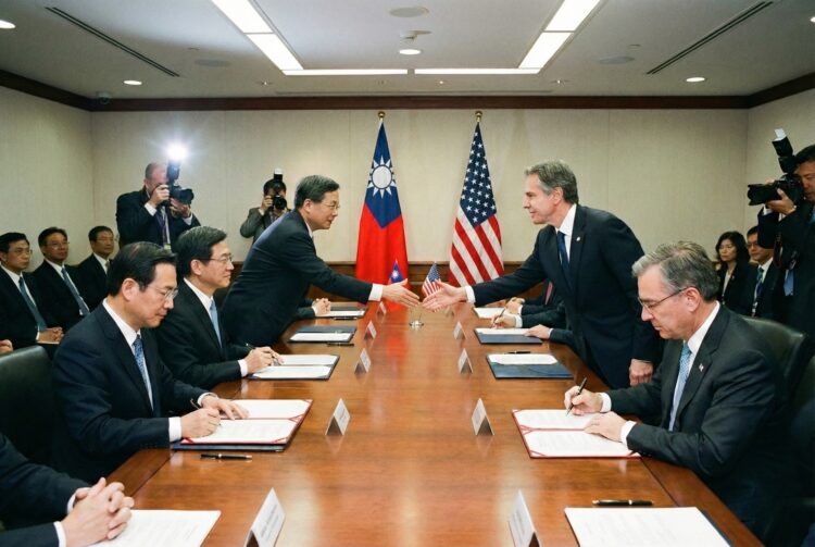 Una fotografía de una sala de conferencias formal muestra a un alto funcionario estadounidense (a la derecha, parecido a Antony Blinken) estrechando la mano de un alto funcionario taiwanés (a la izquierda) sobre una larga mesa de madera pulida. Ambos han estado firmando documentos abiertos frente a ellos. Las banderas de Estados Unidos y de la República de China (Taiwán) están colocadas verticalmente detrás de ellos. Otros funcionarios observan sentados a la mesa, mientras fotógrafos con cámaras y flashes trabajan en el fondo.