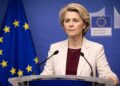 Ursula von der Leyen habla desde un podio en una conferencia de prensa, con expresión firme y seria, micrófonos al frente y el emblema y la bandera de la Unión Europea visibles en el fondo azul institucional.