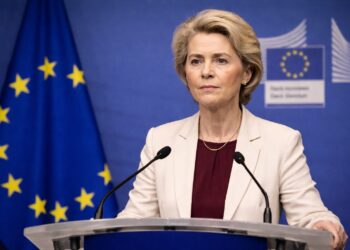 Ursula von der Leyen habla desde un podio en una conferencia de prensa, con expresión firme y seria, micrófonos al frente y el emblema y la bandera de la Unión Europea visibles en el fondo azul institucional.