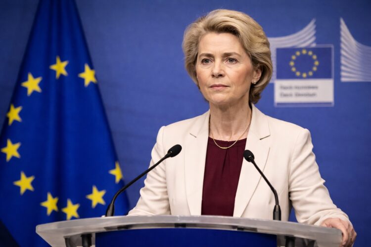 Ursula von der Leyen habla desde un podio en una conferencia de prensa, con expresión firme y seria, micrófonos al frente y el emblema y la bandera de la Unión Europea visibles en el fondo azul institucional.