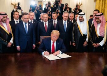 El expresidente de los Estados Unidos, Donald Trump, con traje azul y corbata roja, está sentado en una gran mesa de madera, firmando un documento. Detrás de él, de pie, hay un grupo de hombres, algunos con trajes occidentales y otros con vestimenta tradicional árabe. Al fondo, numerosos fotógrafos con cámaras y flashes capturan el momento en una sala ornamentada.