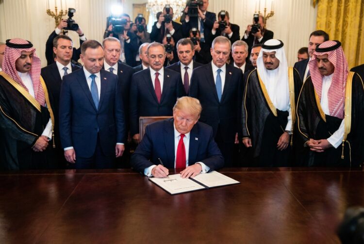 El expresidente de los Estados Unidos, Donald Trump, con traje azul y corbata roja, está sentado en una gran mesa de madera, firmando un documento. Detrás de él, de pie, hay un grupo de hombres, algunos con trajes occidentales y otros con vestimenta tradicional árabe. Al fondo, numerosos fotógrafos con cámaras y flashes capturan el momento en una sala ornamentada.