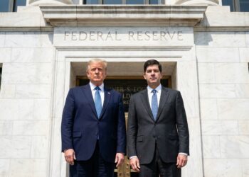 Donald Trump, a la izquierda, y Kevin Warsh, a la derecha, ambos vestidos con trajes oscuros y corbatas azules, posan de pie y mirando a la cámara frente a la entrada de piedra del edificio de la Reserva Federal de los Estados Unidos. El texto "FEDERAL RESERVE" está grabado sobre la puerta dorada. La imagen es una fotografía al aire libre en un día soleado.