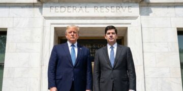 Donald Trump, a la izquierda, y Kevin Warsh, a la derecha, ambos vestidos con trajes oscuros y corbatas azules, posan de pie y mirando a la cámara frente a la entrada de piedra del edificio de la Reserva Federal de los Estados Unidos. El texto "FEDERAL RESERVE" está grabado sobre la puerta dorada. La imagen es una fotografía al aire libre en un día soleado.