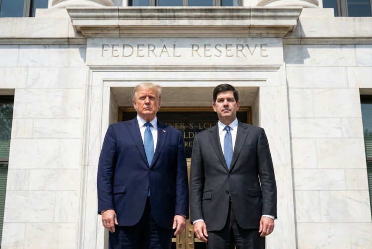 Donald Trump, a la izquierda, y Kevin Warsh, a la derecha, ambos vestidos con trajes oscuros y corbatas azules, posan de pie y mirando a la cámara frente a la entrada de piedra del edificio de la Reserva Federal de los Estados Unidos. El texto "FEDERAL RESERVE" está grabado sobre la puerta dorada. La imagen es una fotografía al aire libre en un día soleado.