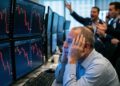 Un hombre de mediana edad con una camisa azul se agarra la cabeza con ambas manos, con los ojos abiertos en una expresión de incredulidad y estrés extremo. Está sentado frente a varios monitores de computadora que muestran gráficos financieros con fuertes caídas marcadas en rojo. En el fondo desenfocado, otros hombres gesticulan y gritan, sugiriendo un caos generalizado en una sala de operaciones bursátiles durante una crisis financiera.