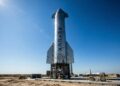 Fotografía a plena luz del día del enorme cohete Starship de acero inoxidable en su plataforma de lanzamiento en las instalaciones de Starbase en Boca Chica, Texas. El nombre "SPACEX" es claramente visible verticalmente en el fuselaje plateado, representando el activo tecnológico clave y la infraestructura de la compañía aeroespacial.