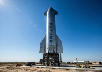 Fotografía a plena luz del día del enorme cohete Starship de acero inoxidable en su plataforma de lanzamiento en las instalaciones de Starbase en Boca Chica, Texas. El nombre "SPACEX" es claramente visible verticalmente en el fuselaje plateado, representando el activo tecnológico clave y la infraestructura de la compañía aeroespacial.