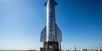 Fotografía a plena luz del día del enorme cohete Starship de acero inoxidable en su plataforma de lanzamiento en las instalaciones de Starbase en Boca Chica, Texas. El nombre "SPACEX" es claramente visible verticalmente en el fuselaje plateado, representando el activo tecnológico clave y la infraestructura de la compañía aeroespacial.
