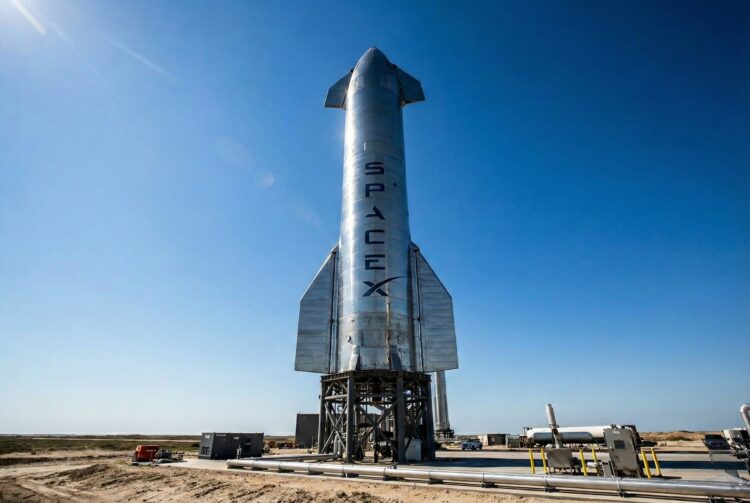 Fotografía a plena luz del día del enorme cohete Starship de acero inoxidable en su plataforma de lanzamiento en las instalaciones de Starbase en Boca Chica, Texas. El nombre "SPACEX" es claramente visible verticalmente en el fuselaje plateado, representando el activo tecnológico clave y la infraestructura de la compañía aeroespacial.