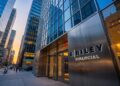 Fotografía de ángulo contrapicado de la entrada moderna de las oficinas de B. Riley Financial en un entorno urbano al atardecer. Un gran letrero de metal cepillado con letras azules iluminadas que dicen "B. RILEY FINANCIAL" destaca junto a las puertas de cristal. Rascacielos de vidrio circundantes y el tráfico de la calle (incluyendo taxis amarillos) llenan el fondo bajo un cielo con tonos naranjas y azules profundos.