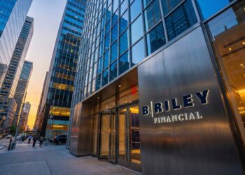 Fotografía de ángulo contrapicado de la entrada moderna de las oficinas de B. Riley Financial en un entorno urbano al atardecer. Un gran letrero de metal cepillado con letras azules iluminadas que dicen "B. RILEY FINANCIAL" destaca junto a las puertas de cristal. Rascacielos de vidrio circundantes y el tráfico de la calle (incluyendo taxis amarillos) llenan el fondo bajo un cielo con tonos naranjas y azules profundos.