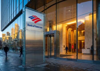 Fotografía exterior al atardecer de la entrada moderna de un edificio de oficinas de Bank of America. Un gran letrero vertical de acero inoxidable con el logotipo rojo y azul y el texto "Bank of America" destaca en primer plano. La fachada de vidrio refleja los rascacielos circundantes y el cielo azul, mientras que el interior del vestíbulo brilla con una luz dorada y cálida. Varios peatones caminan por la acera mojada frente al edificio.