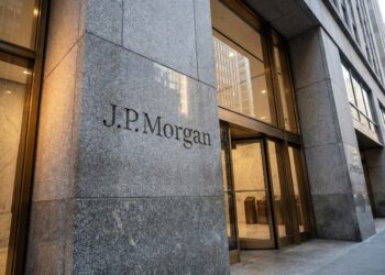 Fotografía exterior en ángulo contrapicado que muestra la entrada de un edificio de oficinas de J.P. Morgan en una ciudad. El nombre "J.P. Morgan" está grabado prominentemente en un gran bloque de granito gris texturizado en el lado izquierdo. A la derecha, unas puertas giratorias de cristal con marcos de metal color bronce reflejan los edificios circundantes y dan acceso a un vestíbulo interior cálidamente iluminado.