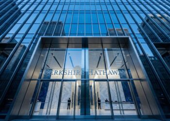 Una fotografía de ángulo contrapicado muestra la moderna fachada de vidrio y metal de la entrada principal del edificio de Berkshire Hathaway. El nombre de la empresa, "BERKSHIRE HATHAWAY", está visible sobre las puertas de acceso. El cielo azul y otros rascacielos se reflejan en los paneles de vidrio de la estructura.