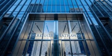 Una fotografía de ángulo contrapicado muestra la moderna fachada de vidrio y metal de la entrada principal del edificio de Berkshire Hathaway. El nombre de la empresa, "BERKSHIRE HATHAWAY", está visible sobre las puertas de acceso. El cielo azul y otros rascacielos se reflejan en los paneles de vidrio de la estructura.