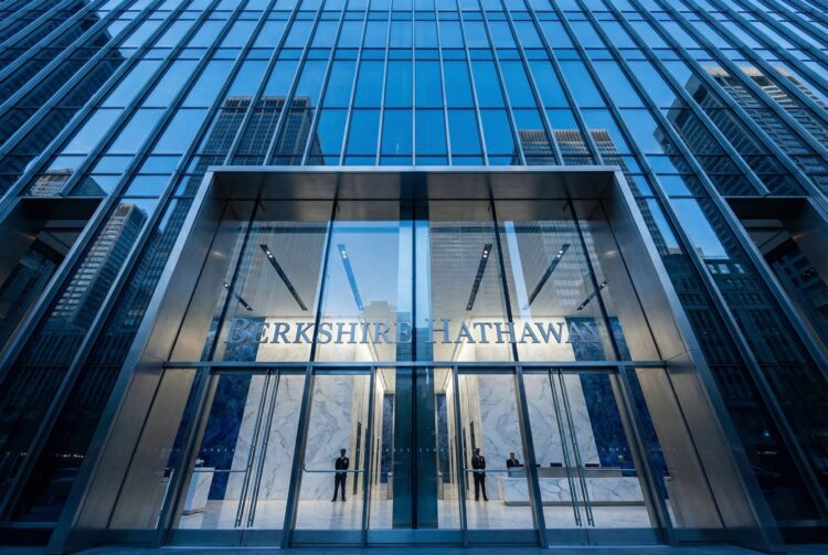 Una fotografía de ángulo contrapicado muestra la moderna fachada de vidrio y metal de la entrada principal del edificio de Berkshire Hathaway. El nombre de la empresa, "BERKSHIRE HATHAWAY", está visible sobre las puertas de acceso. El cielo azul y otros rascacielos se reflejan en los paneles de vidrio de la estructura.
