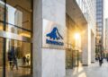 Fotografía exterior de la entrada de la sede de la firma de gestión de inversiones Invesco. El logo azul de la compañía está montado sobre una pared de mármol blanco y la luz del sol poniente se refleja en los ventanales de vidrio del edificio, creando un destello dorado. Se ve gente caminando por la acera y entrando al edificio en un entorno urbano financiero.