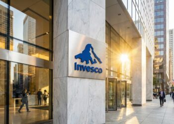 Fotografía exterior de la entrada de la sede de la firma de gestión de inversiones Invesco. El logo azul de la compañía está montado sobre una pared de mármol blanco y la luz del sol poniente se refleja en los ventanales de vidrio del edificio, creando un destello dorado. Se ve gente caminando por la acera y entrando al edificio en un entorno urbano financiero.