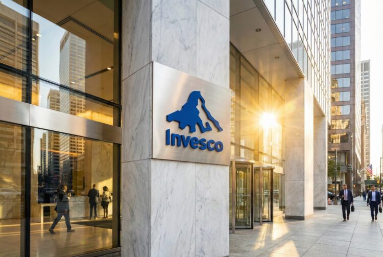 Fotografía exterior de la entrada de la sede de la firma de gestión de inversiones Invesco. El logo azul de la compañía está montado sobre una pared de mármol blanco y la luz del sol poniente se refleja en los ventanales de vidrio del edificio, creando un destello dorado. Se ve gente caminando por la acera y entrando al edificio en un entorno urbano financiero.