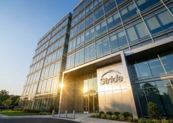 Fotografía exterior en ángulo contrapicado de la entrada principal del edificio de oficinas moderno de la empresa Stride. El logo "Stride" está montado en un panel metálico plateado sobre las puertas de vidrio. La luz dorada del sol poniente golpea la fachada de vidrio, creando un destello brillante. El entorno incluye áreas verdes y un acceso vehicular pavimentado bajo un cielo azul despejado.