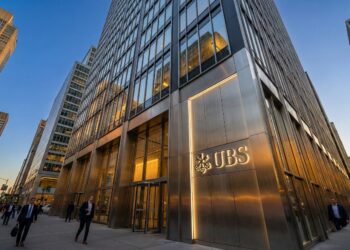 Fotografía exterior del moderno edificio de oficinas de UBS en una ciudad al atardecer. La entrada principal y el gran letrero con el logo de UBS y las llaves están iluminados. La fachada de vidrio y metal refleja la luz del cielo azul y las luces de la ciudad. Varias personas en traje de negocios caminan por la acera frente al edificio.