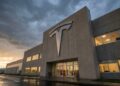 Fotografía exterior en ángulo contrapicado de la masiva entrada de la fábrica Gigafactory de Tesla. Un enorme logotipo "T" de Tesla fabricado en metal cepillado está montado sobre la fachada de hormigón y las puertas de vidrio. El cielo está cubierto de densas nubes grises de tormenta al atardecer, mientras que las luces cálidas del interior de las oficinas brillan y se reflejan en el pavimento mojado exterior.