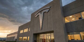 Fotografía exterior en ángulo contrapicado de la masiva entrada de la fábrica Gigafactory de Tesla. Un enorme logotipo "T" de Tesla fabricado en metal cepillado está montado sobre la fachada de hormigón y las puertas de vidrio. El cielo está cubierto de densas nubes grises de tormenta al atardecer, mientras que las luces cálidas del interior de las oficinas brillan y se reflejan en el pavimento mojado exterior.