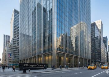 Una fotografía exterior de día muestra la moderna torre de oficinas de vidrio y acero que sirve como sede de JPMorgan Chase en la ciudad de Nueva York. Un gran letrero oscuro a nivel de la calle en la esquina del edificio dice "JPMorgan Chase". Peatones caminan por las aceras y taxis amarillos circulan por la calle concurrida bajo un cielo azul despejado. El edificio refleja los rascacielos circundantes.