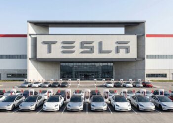 Fotografía industrial de plano general que muestra la entrada principal de una Tesla Gigafactory bajo luz solar directa. La fachada de hormigón y acero presenta el logo de "TESLA" integrado en gran escala. Frente al edificio, numerosos vehículos eléctricos Tesla Model 3 y Model Y están estacionados ordenadamente en filas de Supercargadores. La imagen destaca la magnitud de la instalación manufacturera y la infraestructura de soporte.