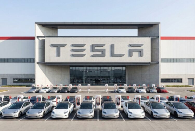 Fotografía industrial de plano general que muestra la entrada principal de una Tesla Gigafactory bajo luz solar directa. La fachada de hormigón y acero presenta el logo de "TESLA" integrado en gran escala. Frente al edificio, numerosos vehículos eléctricos Tesla Model 3 y Model Y están estacionados ordenadamente en filas de Supercargadores. La imagen destaca la magnitud de la instalación manufacturera y la infraestructura de soporte.