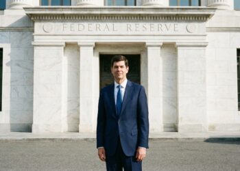 Kevin Warsh, de pie y de frente, vestido con un traje azul marino y corbata azul, posa ante la entrada principal de mármol blanco del edificio de la Reserva Federal en Washington D.C. El texto "FEDERAL RESERVE" está grabado en el dintel de piedra sobre la puerta de bronce. La iluminación es de día.
