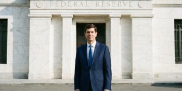 Kevin Warsh, de pie y de frente, vestido con un traje azul marino y corbata azul, posa ante la entrada principal de mármol blanco del edificio de la Reserva Federal en Washington D.C. El texto "FEDERAL RESERVE" está grabado en el dintel de piedra sobre la puerta de bronce. La iluminación es de día.