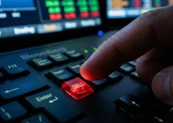 Fotografía de primer plano detalle que muestra el dedo de un operador financiero presionando una tecla roja personalizada e iluminada con la palabra "SELL" (Vender) en un teclado profesional Bloomberg. Al fondo, se perciben pantallas desenfocadas con gráficos y datos bursátiles en tiempo real, reflejando la tensión del trading.