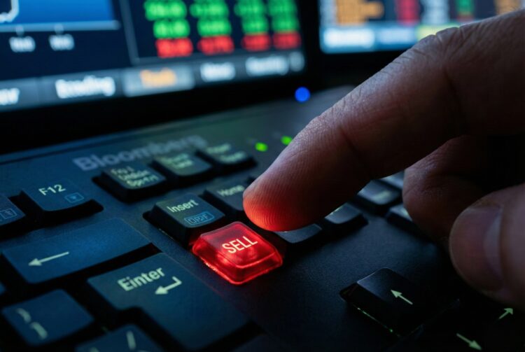 Fotografía de primer plano detalle que muestra el dedo de un operador financiero presionando una tecla roja personalizada e iluminada con la palabra "SELL" (Vender) en un teclado profesional Bloomberg. Al fondo, se perciben pantallas desenfocadas con gráficos y datos bursátiles en tiempo real, reflejando la tensión del trading.