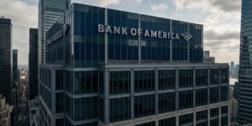 Una fotografía de arquitectura muestra la parte superior de un rascacielos de oficinas en un entorno urbano denso. El letrero con el nombre y el logotipo de "BANK OF AMERICA" es prominente en la fachada de vidrio azul y acero. El edificio está rodeado por otros rascacielos bajo un cielo mayormente nublado con luz natural difusa; un río es visible en el fondo lejano a la derecha.