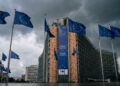 Una fotografía exterior muestra el edificio Berlaymont, sede de la Comisión Europea en Bruselas, bajo un cielo dramático y oscuro lleno de nubes de tormenta. Múltiples banderas azules de la Unión Europea con estrellas amarillas ondean en mástiles frente a la fachada curva de vidrio y acero. Una gran pancarta vertical azul en el edificio dice "European Commission" en varios idiomas. El suelo de la plaza en primer plano está mojado y refleja la luz tenue; no hay personas visibles en la escena.
