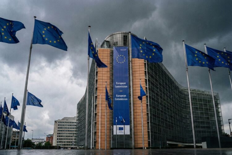 Una fotografía exterior muestra el edificio Berlaymont, sede de la Comisión Europea en Bruselas, bajo un cielo dramático y oscuro lleno de nubes de tormenta. Múltiples banderas azules de la Unión Europea con estrellas amarillas ondean en mástiles frente a la fachada curva de vidrio y acero. Una gran pancarta vertical azul en el edificio dice "European Commission" en varios idiomas. El suelo de la plaza en primer plano está mojado y refleja la luz tenue; no hay personas visibles en la escena.