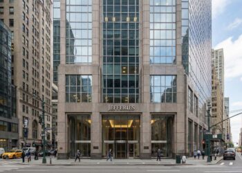 Fotografía exterior a nivel de calle de la entrada principal de la sede de la firma de inversión Jefferies en 520 Madison Avenue, Nueva York. La fachada sólida de granito y vidrio muestra un letrero prominente con el nombre "JEFFERIES" sobre las puertas giratorias. Peatones caminan por la acera en un día claro en Manhattan.