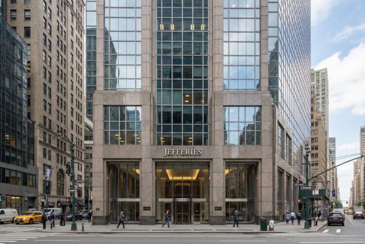 Fotografía exterior a nivel de calle de la entrada principal de la sede de la firma de inversión Jefferies en 520 Madison Avenue, Nueva York. La fachada sólida de granito y vidrio muestra un letrero prominente con el nombre "JEFFERIES" sobre las puertas giratorias. Peatones caminan por la acera en un día claro en Manhattan.