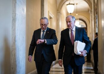 Fotografía de los senadores Chuck Schumer (izquierda), mirando su reloj con preocupación, y otro senador (derecha) sosteniendo un fajo de documentos, mientras caminan con urgencia por un pasillo de mármol del Capitolio de los Estados Unidos. La iluminación es cálida y la atmósfera parece tensa, sugiriendo la proximidad de una fecha límite importante o una votación inminente.