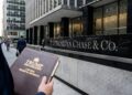 Una fotografía a la altura de los ojos muestra a una persona, vista desde el torso hacia abajo, sosteniendo una carpeta de cuero marrón con el texto dorado "TRUMP ORGANIZATION" y "LAW FILE: TRUMP VS. JPMORGAN". La persona está de pie frente a un edificio de oficinas moderno, con un gran letrero plateado que dice "JPMORGAN CHASE & CO." visible en una pared de granito oscuro en el fondo. Hay peatones caminando por la acera y la entrada del edificio al fondo.