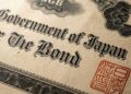 Fotografía macro de un documento de papel envejecido y texturizado que muestra el texto "Government of Japan" y "The Bond" en una tipografía negra ornamentada. Un sello oficial rojo cuadrado con caracteres japoneses es visible en la esquina inferior derecha, rodeado de patrones de grabado detallados.